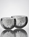 Two clear glass art objects with embedded air bubbles, displayed on a reflective surface against a light gray background.