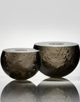 Two spherical smoked glass art objects with a textured, bubble-like surface and a smooth white rim, displayed on a reflective surface against a light gray background.