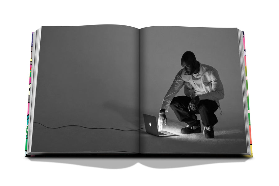 Assouline Louis Vuitton: Virgil Abloh Classic Cartoon Cover coffee table book displaying Grey Image of Virgil Abloh using Apple Mac Laptop on a white back ground available at Spacio India for luxury home decor accessories collection of Fashion Coffee Table Books.