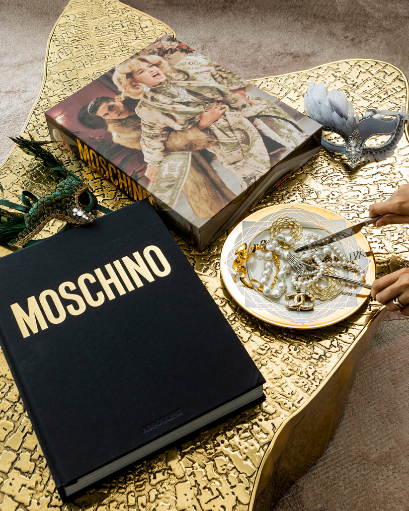 Assouline Moschino coffee table book on a gold finished coffee table with a plate full of Gold & Pearl Jewellery & Masks available at Spacio India for luxury home decor accessories collection of Fashion Coffee Table Books.