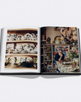 An open hardcover book laid flat on a light gray background, showing two printed interior scenes: shelves of decorative dishes on the left page and a person seated at a table in a shop on the right page.