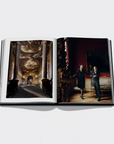 An open hardcover book laid flat on a light gray background, showing two interior scene photographs printed on the pages: a grand classical hall with columns and a staircase on the left, and a dark red room with two people standing on the right.