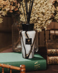 A reed diffuser bottle with a black and white marble pattern, placed on a green book next to stacked vintage books, with dried flowers in the background.