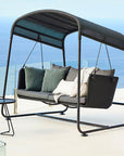 Cave Swing Sofa by Cane-line, designed by Jesper K. Thomsen, featuring a modern canopy design, soft cushions, and weather-resistant materials for indoor and outdoor use. Along with side table on a high rise building balcony in Mumbai