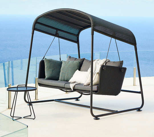 Cave Swing Sofa by Cane-line, designed by Jesper K. Thomsen, featuring a modern canopy design, soft cushions, and weather-resistant materials for indoor and outdoor use. Along with side table on a high rise building balcony in Mumbai