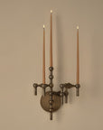 A wall-mounted three-arm candelabra-style light fixture in a dark bronze finish, holding three tall taper candles with visible flames, set against a plain beige wall.