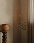 A modern chrome finished metal candelabra with three arms and lit taper candles, placed in a softly lit room corner next to a terracotta pot on a turned wood pedestal.