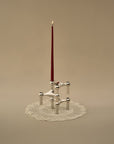A silver-toned metal candle holder with a tall, thin dark red candle, displayed on a white lace doily against a solid beige background.