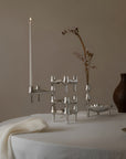 A polished chrome metal candelabra set displayed on a white tablecloth, styled with a tall white taper candle, a dried branch in a clear vase, and a dark brown ceramic jug against a warm beige background.