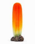 A modern sculpture like a tall, ribbed cactus or flame, with a color gradient from bright orange at the top to pale yellow and light green at the bottom, mounted on a dark brown porous rock base against a solid white background.