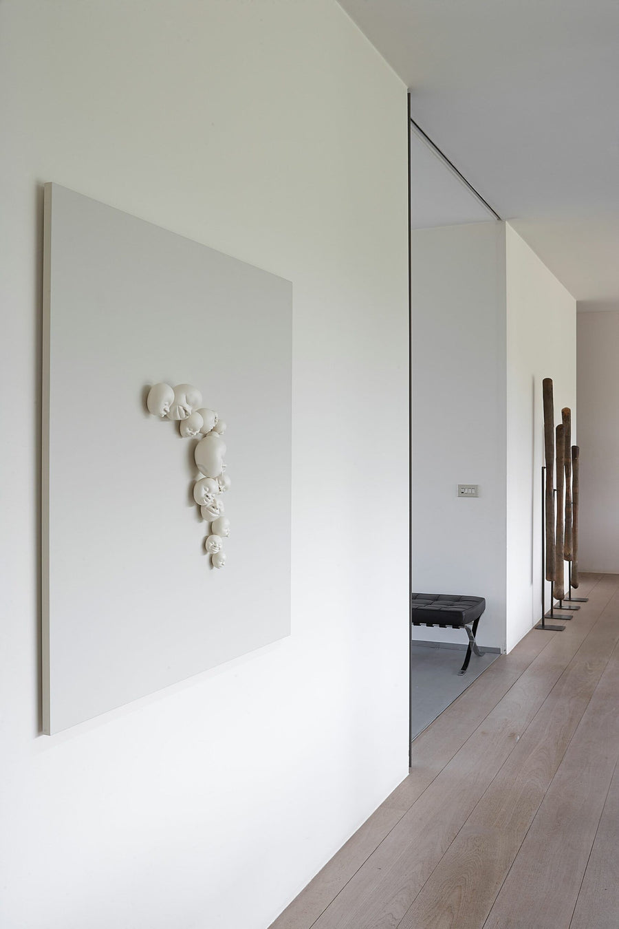 A white wall adorned with a Gardeco Wall Sculpture Happy Faces, created by the talented Brazilian ceramist Selma Calheira. The sculpture features beautiful white ceramic faces and is designed by the brand Gardeco.