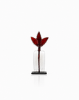 A small decorative glass sculpture of a tulip with a deep red flower and stem, set in a clear cylindrical glass vase on a black base, photographed against a seamless white background.