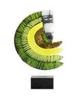 A modern abstract glass sculpture on a clear acrylic stand with a black base, featuring layered green and yellow crescent shapes with a stylized face on a white segment, set against a plain white background.