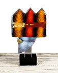 A decorative glass sculpture of a helmet with a crown-like top, featuring orange, red, and gold textured panels, mounted on a silver metal rod over a black rectangular base, set on a light wood surface against a white background.