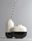 Two modern ceramic vases in a minimalist studio setting. One tall vase is off-white on top with a glossy black base and holds a thin black-and-white striped stick.