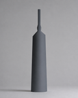 A minimalist, matte dark gray ceramic vase with a flared base and a narrow neck, standing upright against a neutral gray background.