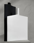 Two modern abstract ceramic vases in matte black and matte white, standing upright on a light gray pedestal against a textured gray concrete wall.