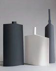 Three minimalist geometric ceramic vases in matte black, white, and dark gray, arranged on a light gray surface against a neutral gray background.