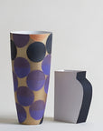 A tall sculptural vase with a gold base and large overlapping purple and navy circles, shown beside a smaller matching vase with a white front and dark navy side on a light gray background.