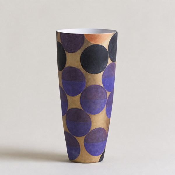 A tall, tapered ceramic vase with a modern abstract pattern of overlapping circles in purple, black, and terracotta on a warm beige background, set against a solid light gray studio backdrop.