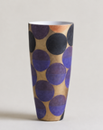 A tall, tapered ceramic vase with a modern abstract pattern of overlapping circles in purple, black, and terracotta on a warm beige background, set against a solid light gray studio backdrop.