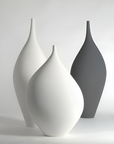 Three matte ceramic vases with a teardrop silhouette and narrow necks, arranged in a studio setting. The front vase is white, the middle vase is partially obscured behind it in white, and the rear vase is dark gray.