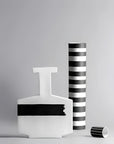 Three modern abstract ceramic vases in a minimalist studio setting against a light gray background. The set includes a white vase with a black horizontal stripe, a tall black-and-white striped vase, and a small black-and-white striped cylinder vase lying on its side.