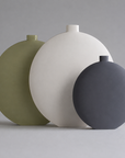 Three matte ceramic vases in olive green, white, and charcoal gray, arranged in a row against a neutral gray background.