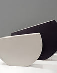 Two modern semi-circular ceramic vases in matte off-white and matte black, displayed against a neutral gray background by Kose Milano, available at Spacio in India. 