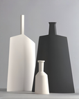 Three minimalist matte ceramic vases in white and charcoal gray, arranged on a light gray surface against a neutral gray background.