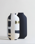 Two modern ceramic vases with a minimalist abstract design, one in white with black and beige geometric blocks and a gold metallic band, and the other in matte black, set against a clean light gray background.