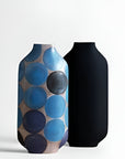 Two modern ceramic vases on a light gray background. The left vase features a pattern of overlapping blue, navy, and gray circles on a beige base, while the right vase is solid matte black.