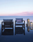 Two chairs and a Les Jardins LJ Solar Lantern SKAAL Tink121 Large 500L Duratek sitting on a concrete slab in front of the ocean create an outdoor oasis with organic charm.