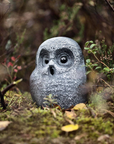 A gray owl-shaped garden statue with a textured stone finish, nestled among green moss, small plants, and forest floor debris.