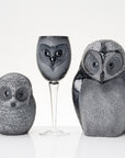 Three owl-shaped decorative objects on a white reflective surface against a white background: a small owl figure, a wine glass with an owl face, and a larger owl figure. The surfaces are mottled black, gray, and white with glossy highlights.