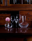 A set of clear glass drinking vessels, including a decanter, a small tumbler, a stemmed wine glass, and a small vase with a pink flower, arranged on a dark wooden cabinet.