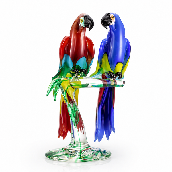 A colorful Murano glass sculpture of two parrots perched on a clear glass branch and base, photographed on a clean white background by Zanetti, available at Spacio in India. 