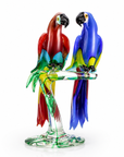 A colorful Murano glass sculpture of two parrots perched on a clear glass branch and base, photographed on a clean white background by Zanetti, available at Spacio in India. 