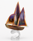 A decorative Murano glass sailboat sculpture with iridescent purple, orange, and gold-toned sails mounted on a clear glass wave-shaped base, photographed against a clean white studio background.