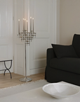 A tall silver floor candelabra with multiple lit taper candles stands in a bright, modern living room next to a black sofa and white walls.