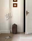 A tall silver metal candelabra with multiple candle holders and lit candles stands in a bright white hallway next to a white door, with a dark brown ceramic vase on the floor.