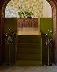 A modern staircase with olive green side panels and a wooden handrail, set within a dark wood arched doorway. Two tall silver candelabra-style floor lamps stand on either side, and a floral arrangement sits above the stairs against a textured light wall.