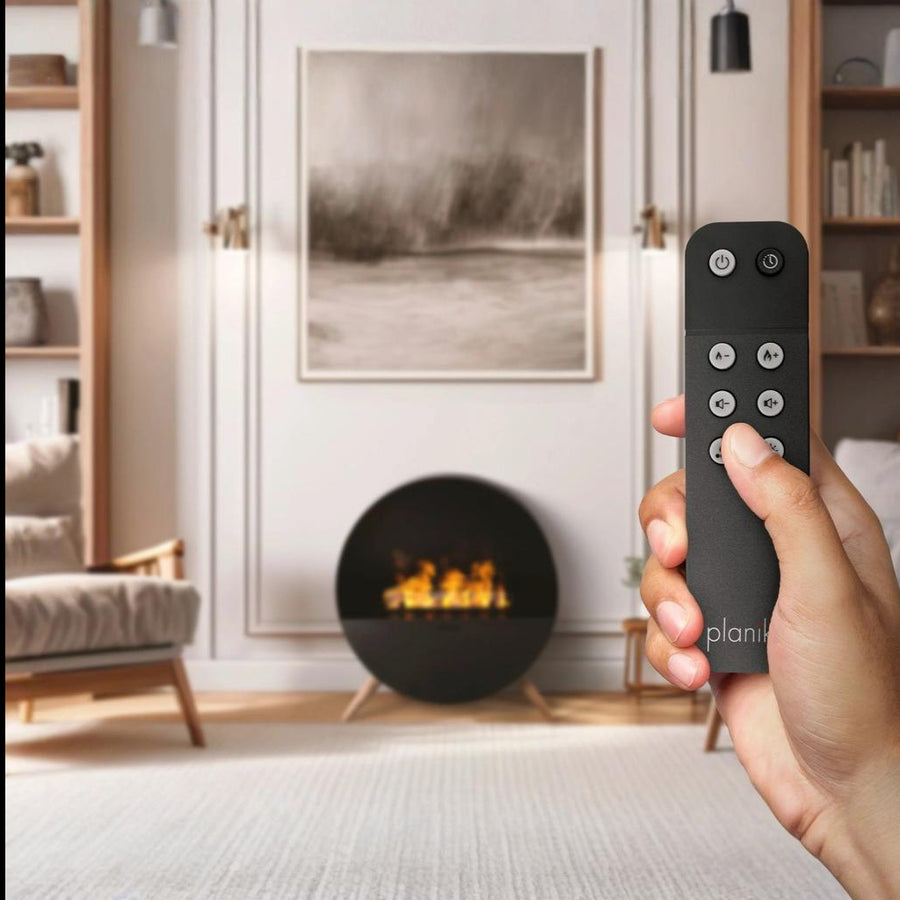 Hand holding a remote control with a modern living room in the background featuring a free standing artificial water vapour fireplace from Planika available at Spacio retail stores in India. 
