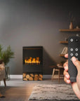 Hand holding a remote control in a cozy living room with a artificial water vapour fireplace by Planika available at Spacio retail stores in India. 