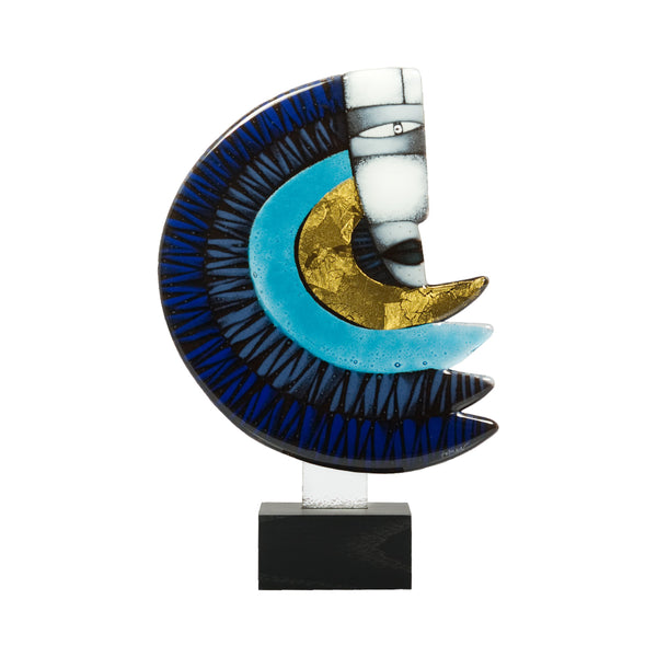 An abstract crescent moon glass sculpture made of layered blue, black, turquoise, white, and gold glass, mounted on a black rectangular base against a plain white background.