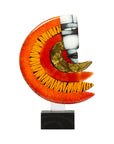 A modern abstract glass sculpture featuring a crescent moon and stylized face motif, mounted on a white vertical bar with a black marble base. The glass has vibrant orange and red gradients, textured black lines, a gold leaf pattern, and a white face with black details, set against a plain white background.