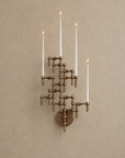 A multi-tiered wall sconce in an antique bronze finish, mounted on a textured beige wall. The fixture features five candle-style lights with white shades and glowing flames.