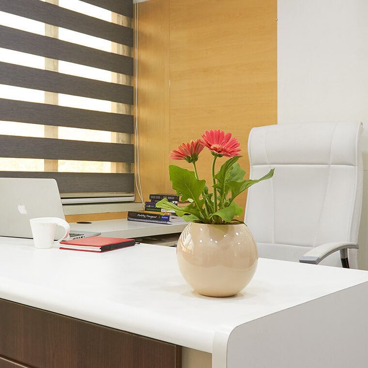 Modern office desk with laptop, books, and a flower vase under striped window blinds.