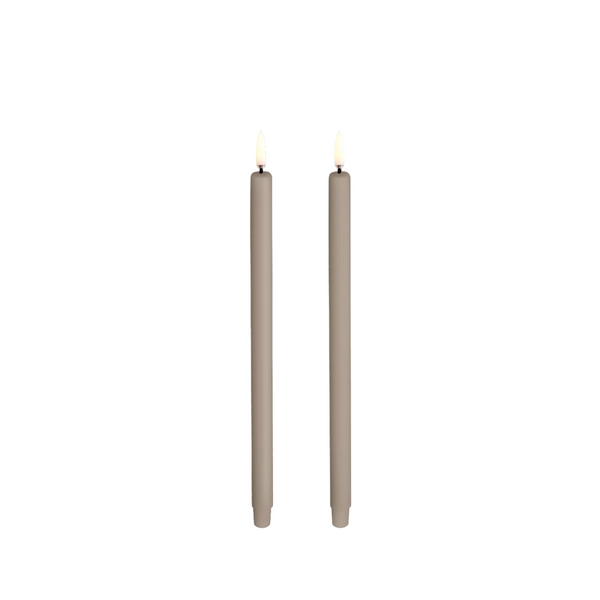 Two tall, mini ulta slim LED taper candles in a warm taupe-beige color, shown lit with small yellow flames against a solid white background from Uyuni available at Spacio in India. 
