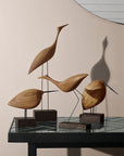 Four Warm Nordic Beak Bird Tall Heron Oak figurines, are elegantly displayed on a table in front of a mirror. The minimalist Danish design perfectly showcases the beauty of natural wood.